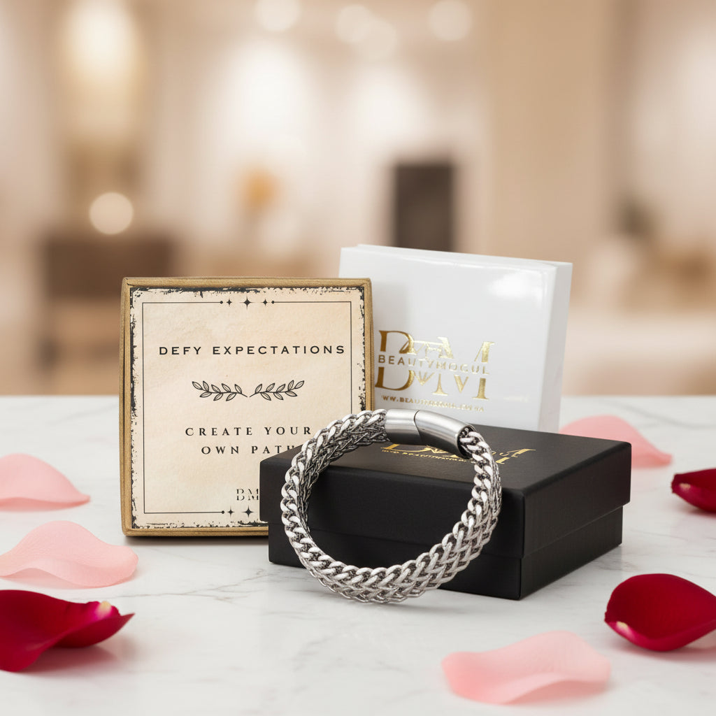 A silver braided chain bracelet displayed on a black gift box, with a cream branded card and a white box behind it on a marble surface scattered with pink and red rose petals.