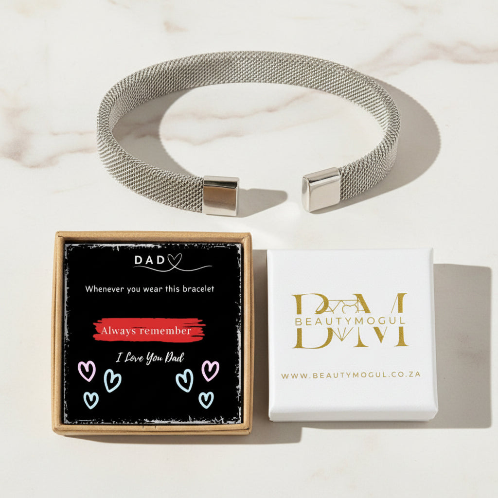A silver mesh bracelet with square metal clasps is displayed on a light marble surface, alongside its gift packaging. One box shows a black insert with a red and white message for Dad, and the other is a white box with gold 'Beautymogul' branding.