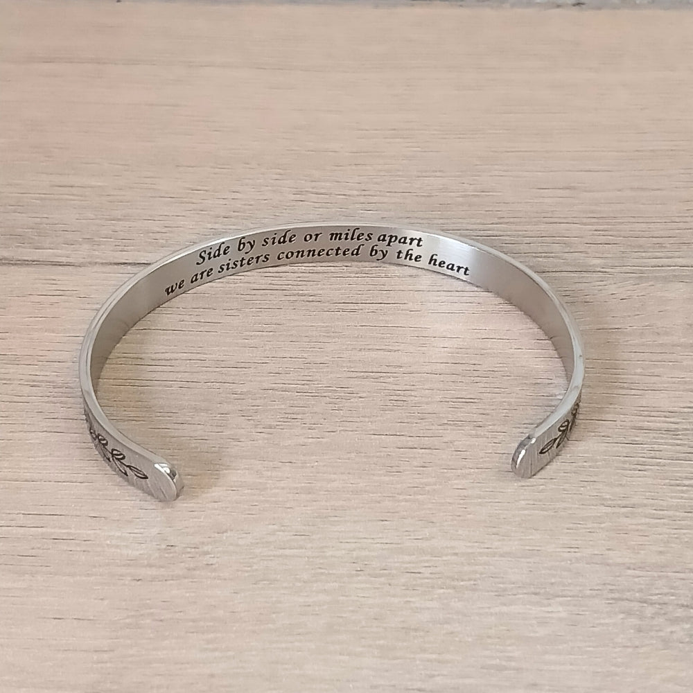 A silver cuff bracelet engraved with the words 'Side by side or miles apart we are sisters connected by the heart' resting on a white marble surface next to eucalyptus leaves.