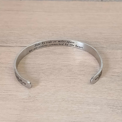 A silver cuff bracelet engraved with the words 'Side by side or miles apart we are sisters connected by the heart' resting on a white marble surface next to eucalyptus leaves.