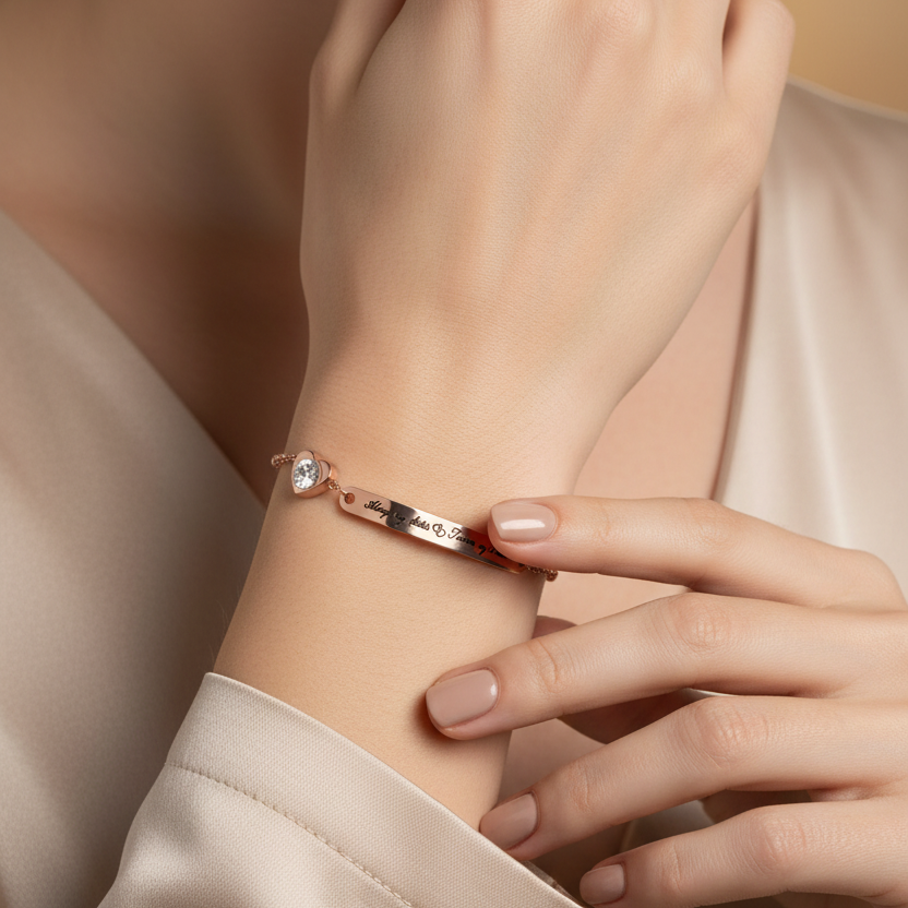 A close-up lifestyle shot of a model wearing a delicate rose gold bracelet with a small clear gemstone and a tiny red accent, styled with a beige satin top.