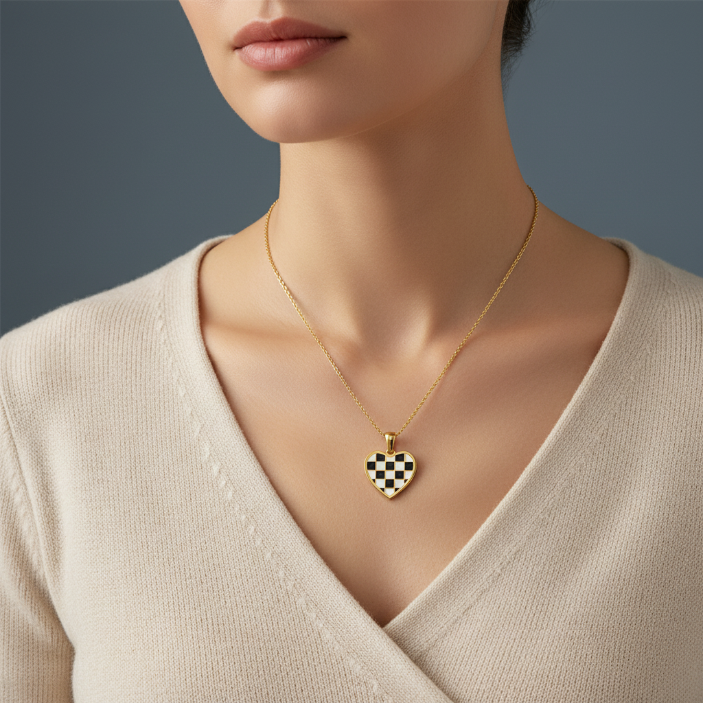 A close-up of a model wearing a delicate gold chain necklace with a heart-shaped pendant featuring a black and white checkerboard pattern, styled with a cream-colored knit top against a dark gray background.