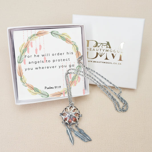 A silver-toned dreamcatcher pendant necklace with feather charms, displayed in front of its white gift box and card. The card features a watercolor wreath and the Bible verse Psalms 91:11.