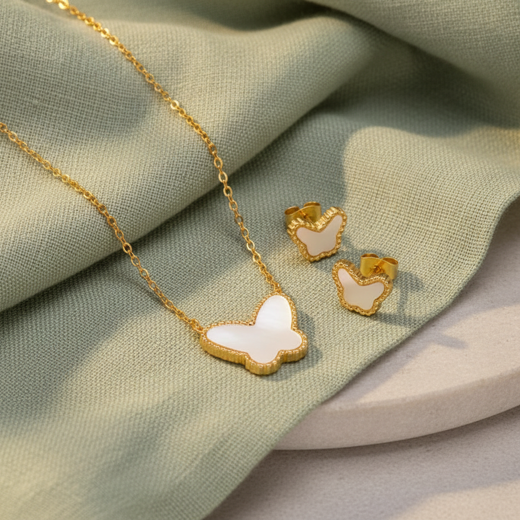 Gold jewelry set featuring a butterfly-shaped pendant necklace with a white inlay and matching butterfly stud earrings, displayed on a warm cream surface.
