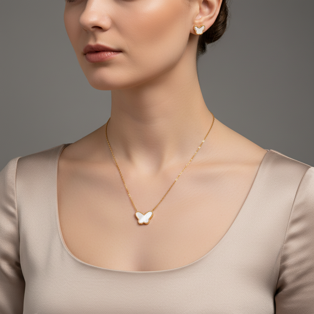 A close-up of a woman wearing a delicate gold necklace with a white butterfly pendant and a matching butterfly stud earring, paired with a beige satin top against a neutral gray background.
