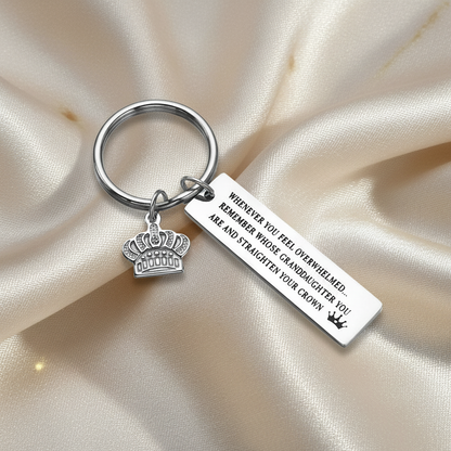 A silver metal keychain with a round key ring, a crown charm, and a rectangular engraved tag reading, 'WHENEVER YOU FEEL OVERWHELMED, REMEMBER WHOSE GRANDDAUGHTER YOU ARE AND STRAIGHTEN YOUR CROWN.' The keychain is photographed on a soft beige satin fabric background.