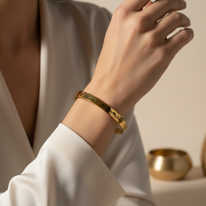 Beautymogul-He-Anchors-My-Soul-Bracelet A close-up of a woman's hand and wrist wearing a polished gold bangle bracelet with engraved characters, set against a warm beige background with decorative gold vases.