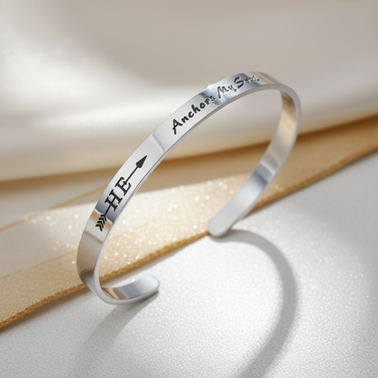 A polished silver cuff bracelet with engraved black lettering, resting on a white textured surface with a gold ribbon. The engraving includes the words '-He-Anchors-My-Soul-