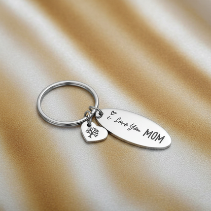 A silver metal keychain with a round key ring, an oval tag engraved with 'I Love You MOM', and a small heart-shaped tag engraved with a tree, resting on a draped beige satin fabric background.