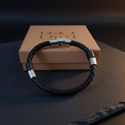 A black braided bracelet with silver-toned metal end caps and a magnetic clasp, displayed in front of a beige branded gift box on a dark surface.