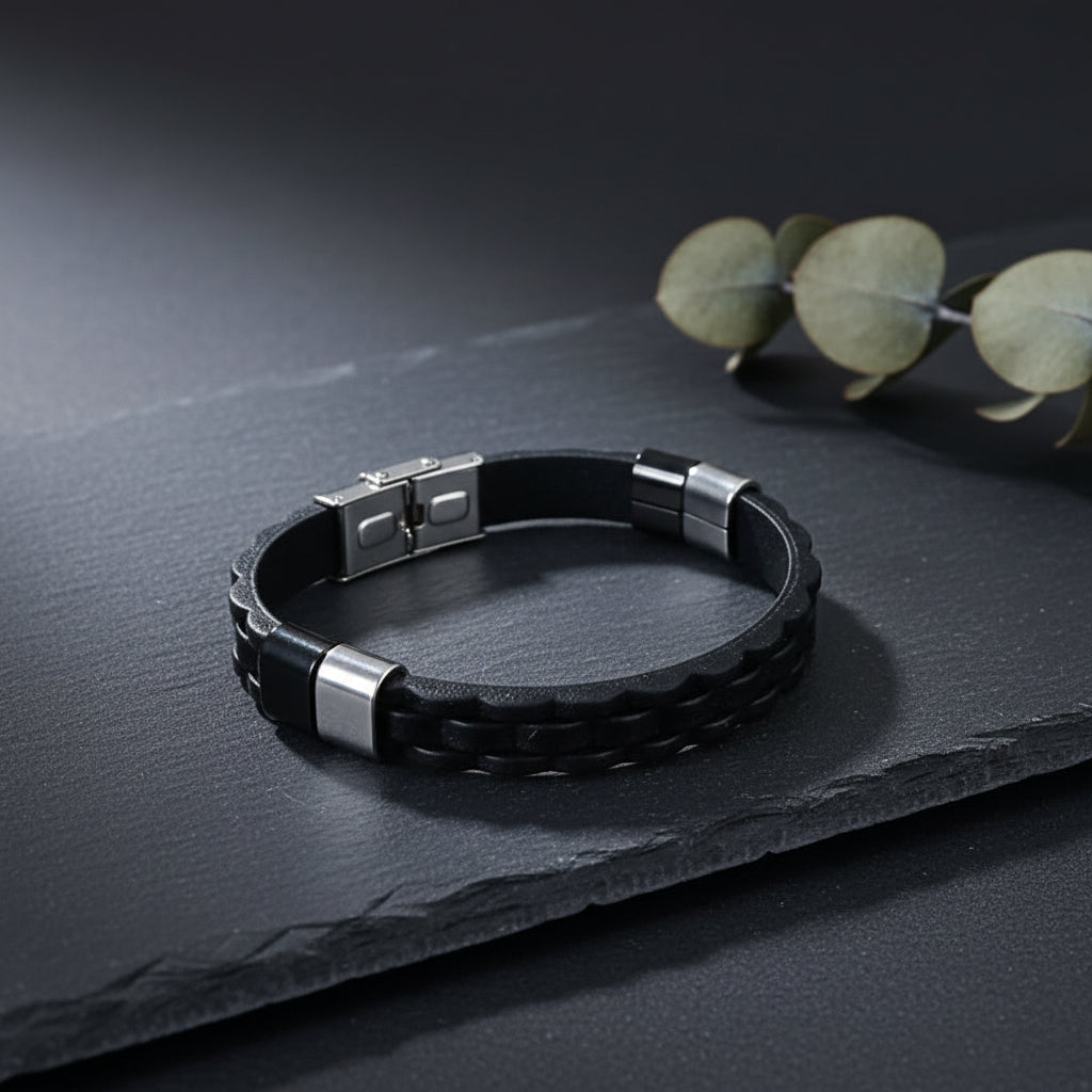 A black braided leather bracelet with silver-toned metal end caps and a square magnetic clasp, displayed on a dark slate surface with eucalyptus leaves in the background.