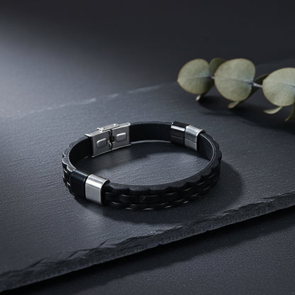 A black braided leather bracelet with silver-toned metal end caps and a square magnetic clasp, displayed on a dark slate surface with eucalyptus leaves in the background.