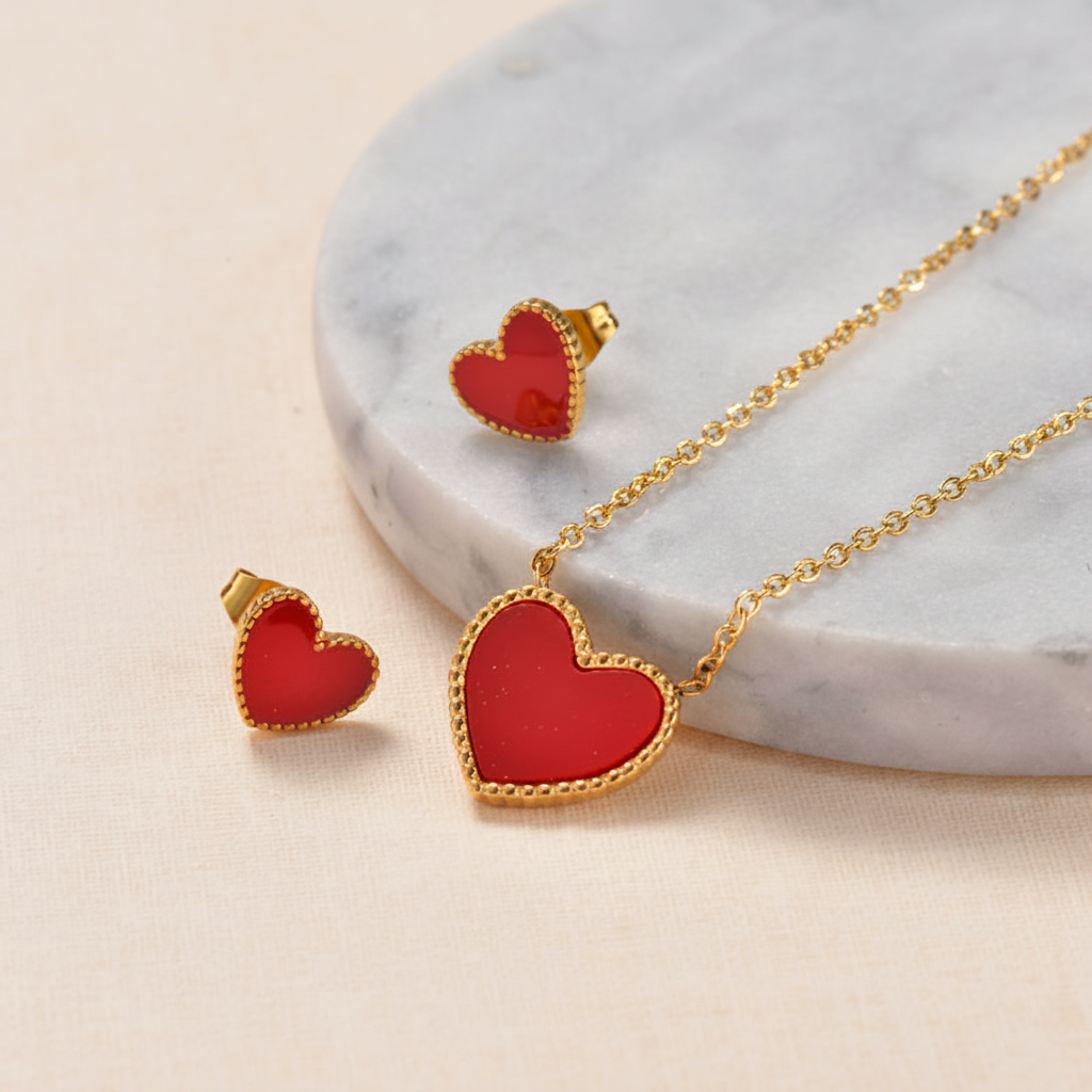 Gold heart jewelry set featuring a pendant necklace and matching stud earrings with red heart inlays, arranged on a soft cream background with gold ribbon accents.