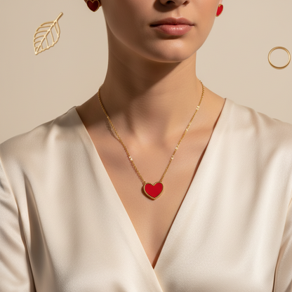 A close-up of a model wearing a delicate gold chain necklace with a red heart-shaped pendant, styled with matching red heart stud earrings and a gold leaf earring against a warm beige background.