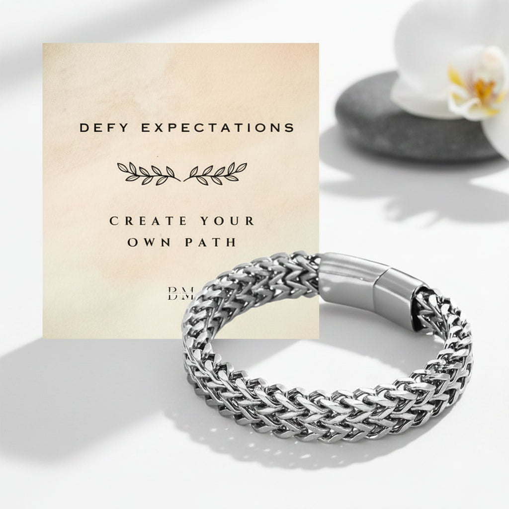A silver braided chain bracelet with a polished clasp, displayed in front of a beige card that reads 'DEFY EXPECTATIONS CREATE YOUR OWN PATH'. The background is a clean white surface with soft shadows and a blurred white flower on a stone.