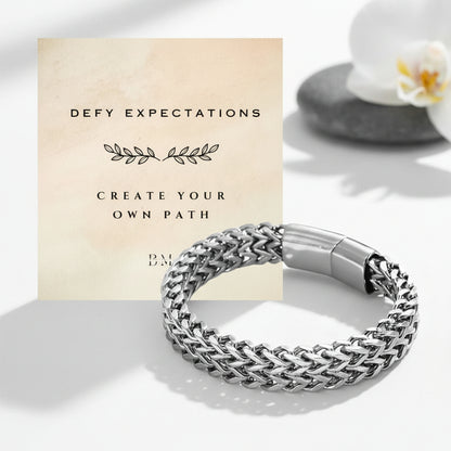 A silver braided chain bracelet with a polished clasp, displayed in front of a beige card that reads 'DEFY EXPECTATIONS CREATE YOUR OWN PATH'. The background is a clean white surface with soft shadows and a blurred white flower on a stone.