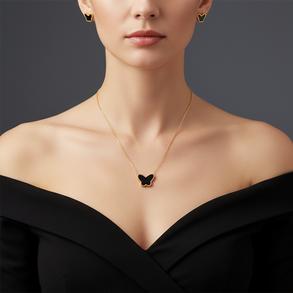 A close-up of a model wearing a delicate gold necklace with a black butterfly pendant, matching gold butterfly stud earrings, and a black off-the-shoulder top against a dark gray background.