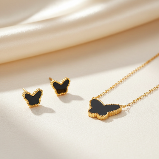 Gold butterfly jewelry set including a necklace and matching stud earrings with black inlay, arranged on a cream satin fabric background.