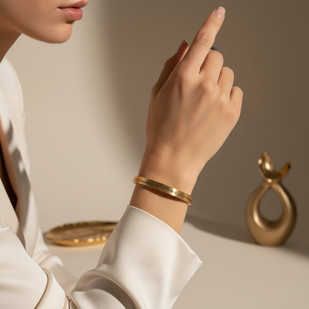 Beautymogul Gold Plated Friendship Journey bracelet- A close-up of a person wearing a slim gold bangle bracelet with engraved text, styled with a white satin blouse against a warm beige background.