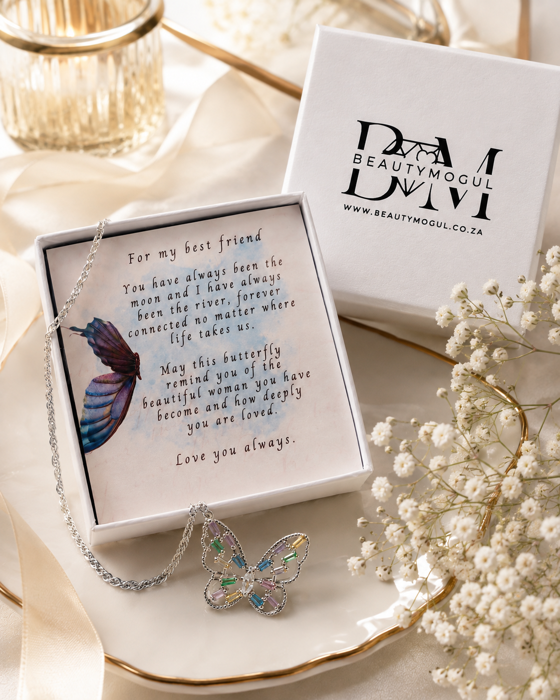 A silver butterfly pendant necklace with iridescent blue and green accents, presented in a white gift box with a sentimental message for a best friend. A second white box with the brand logo 'BEAUTYMOGUL' is visible in the background, styled with cream satin fabric and white baby's breath flowers.