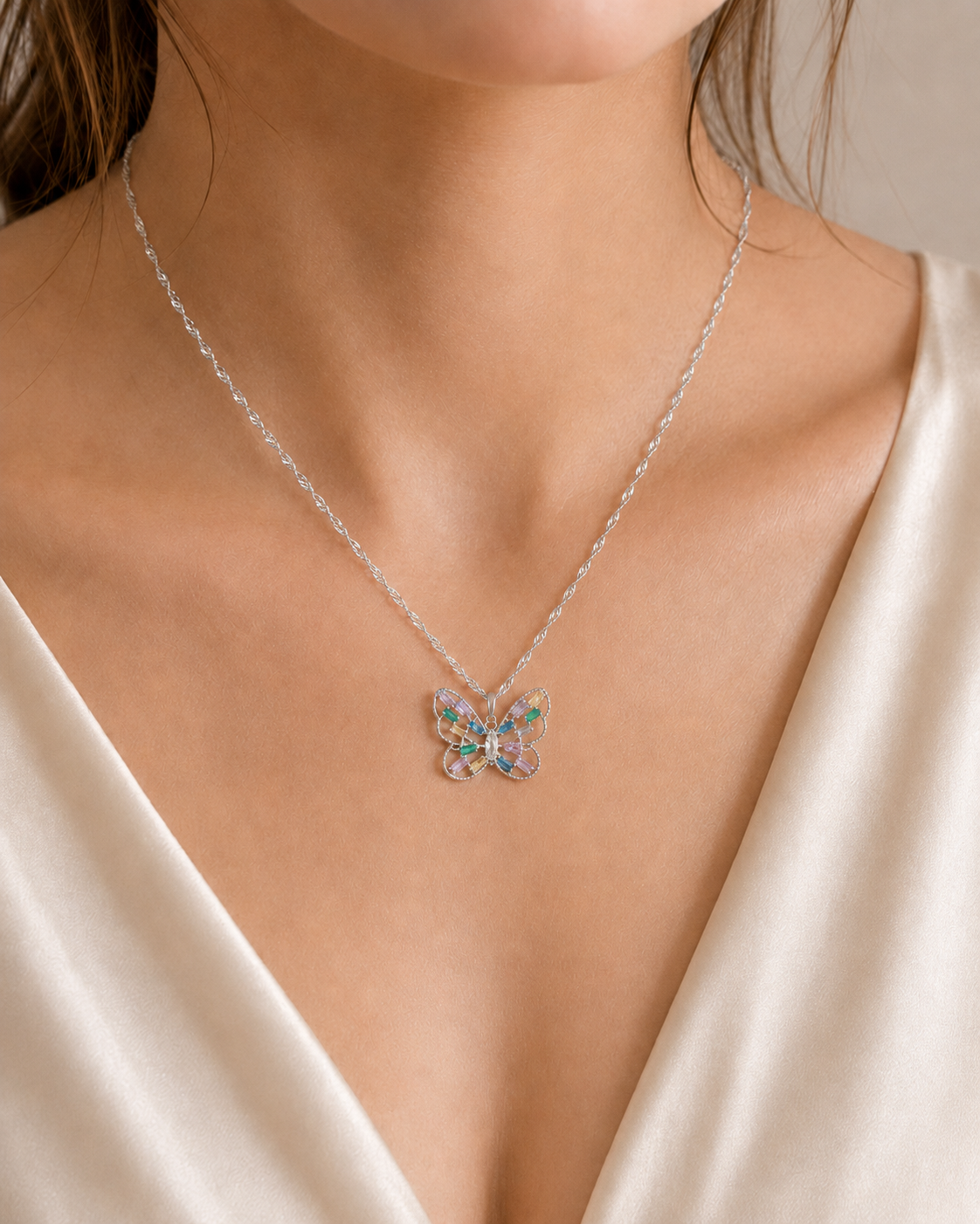 A close-up of a model wearing a delicate silver chain necklace with a butterfly pendant featuring iridescent, multicolored gemstones, styled with a cream satin top.