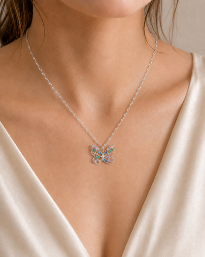 A close-up of a model wearing a delicate silver chain necklace with a butterfly pendant featuring iridescent, multicolored gemstones, styled with a cream satin top.