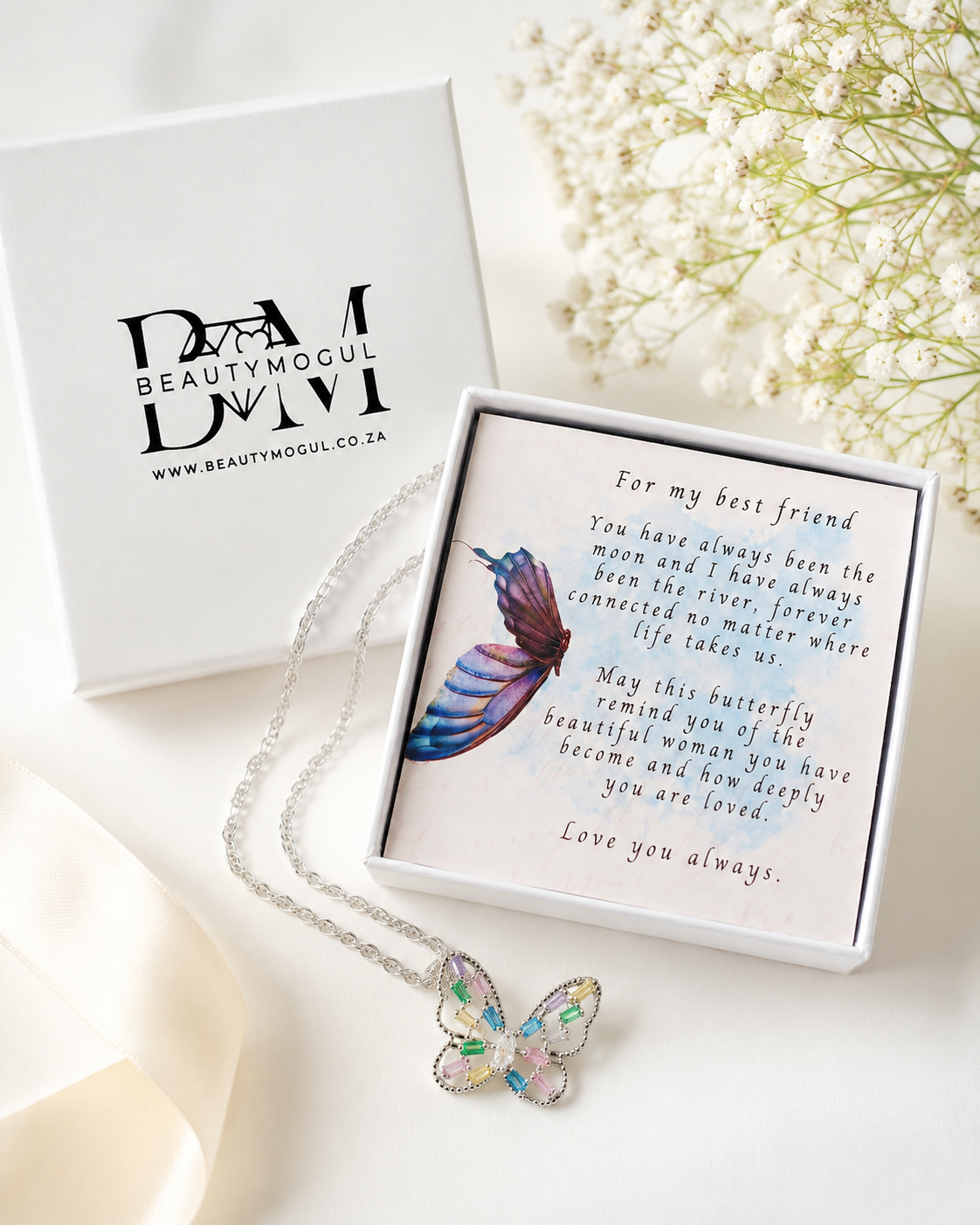 A silver butterfly necklace with iridescent pastel-colored wings is displayed in an open white gift box next to the branded Beautymogul box. The necklace is laid on a soft cream fabric with dried white flowers in the background.