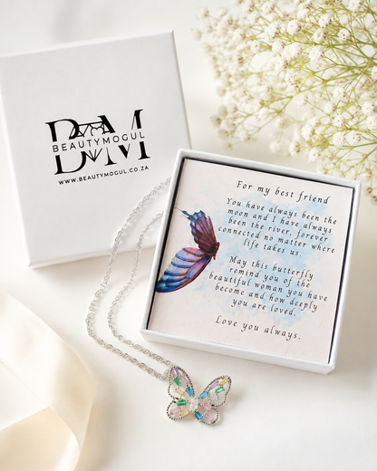 A silver butterfly necklace with iridescent pastel-colored wings is displayed in an open white gift box next to the branded Beautymogul box. The necklace is laid on a soft cream fabric with dried white flowers in the background.