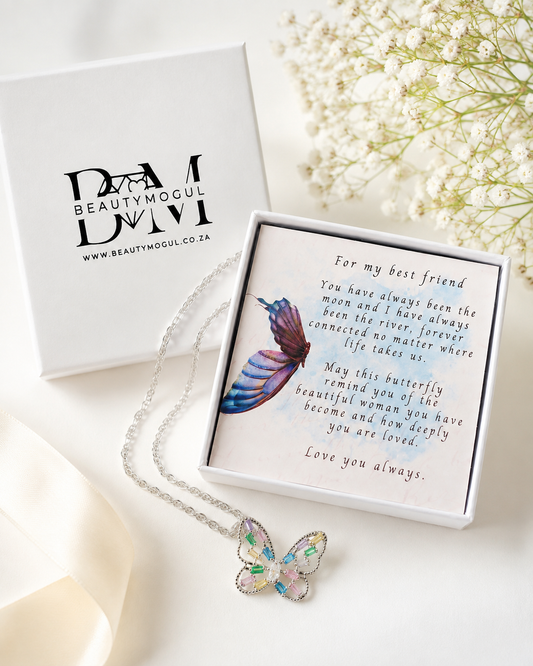 A silver butterfly necklace with iridescent pastel-colored wings is displayed in an open white gift box next to the branded Beautymogul box. The necklace is laid on a soft cream fabric with dried white flowers in the background.