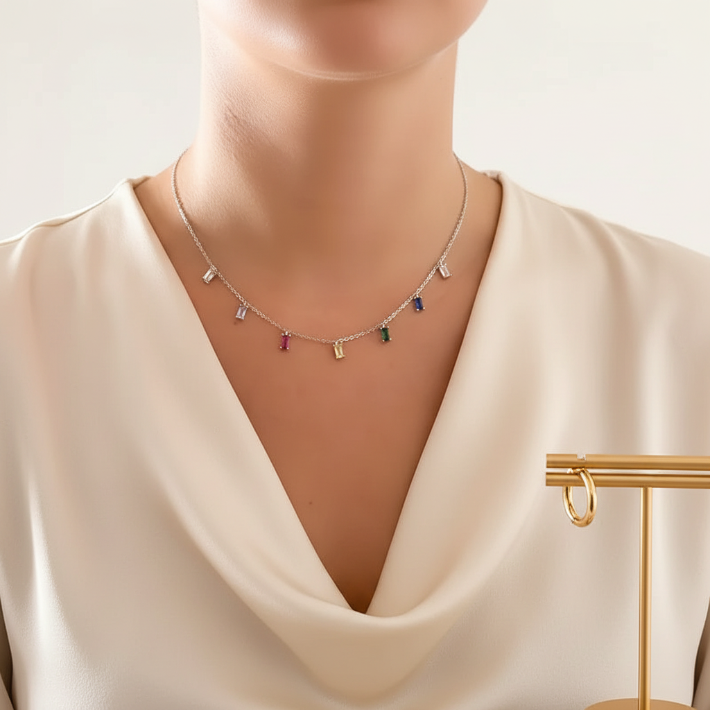 A close-up of a model wearing a delicate silver chain necklace with small colorful rectangular charms, paired with a cream satin top. A gold jewelry display stand is visible on the right side.