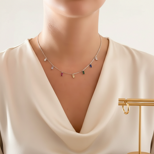 A close-up of a model wearing a delicate silver chain necklace with small colorful rectangular charms, paired with a cream satin top. A gold jewelry display stand is visible on the right side.