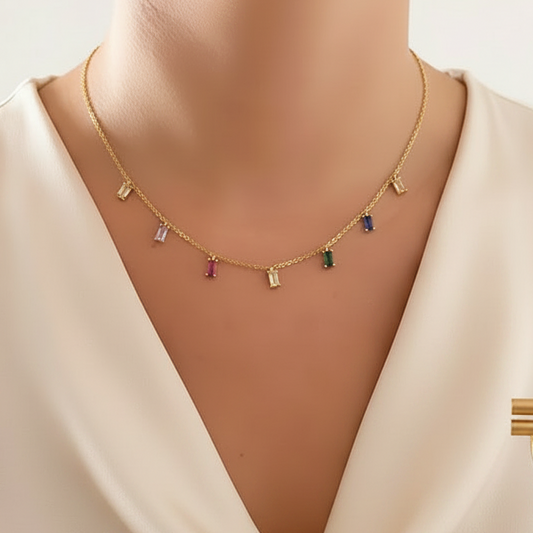A close-up of a model wearing a delicate gold chain necklace with small colorful rectangular gemstone charms, paired with a cream satin cowl-neck top.