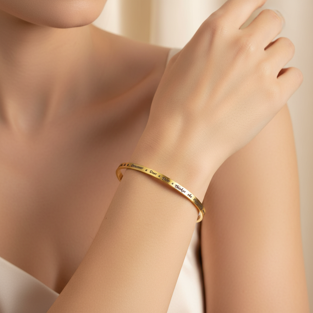 A close-up of a woman's wrist wearing a thin gold bangle bracelet with engraved names, set against a soft beige background.