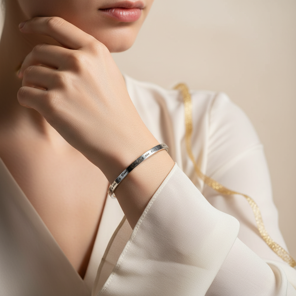 A close-up lifestyle shot of a model wearing a slim silver bangle bracelet with engraved text, paired with a cream-colored satin blouse against a soft beige background.