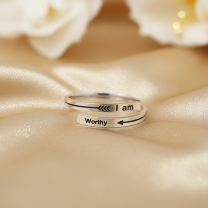 Two silver open-band rings with black engraved text and arrows, one reading 'I am' and the other 'Worthy', displayed on a beige satin fabric with blurred white flowers in the background.