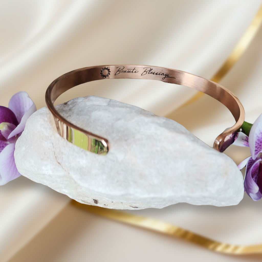 A rose gold cuff bracelet engraved  and a small sunburst logo, resting on a white stone with purple orchids and draped cream fabric in the background.