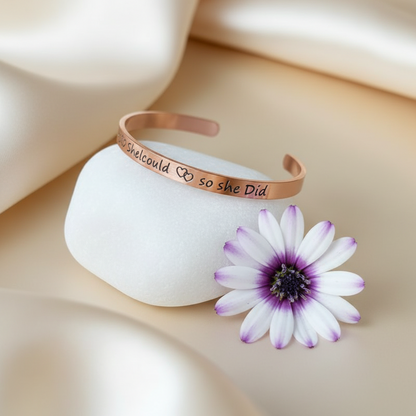 A rose gold cuff bracelet with engraved text and a heart symbol, displayed on a white stone next to a purple and white daisy flower on a beige satin background.