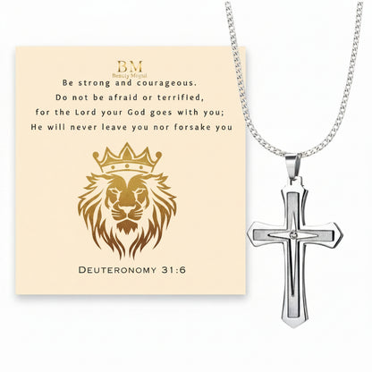 Silver cross pendant necklace displayed next to a cream-colored card featuring a gold lion illustration, the text 'Be strong and courageous' from Deuteronomy 31:6, and a small 'BM' monogram.