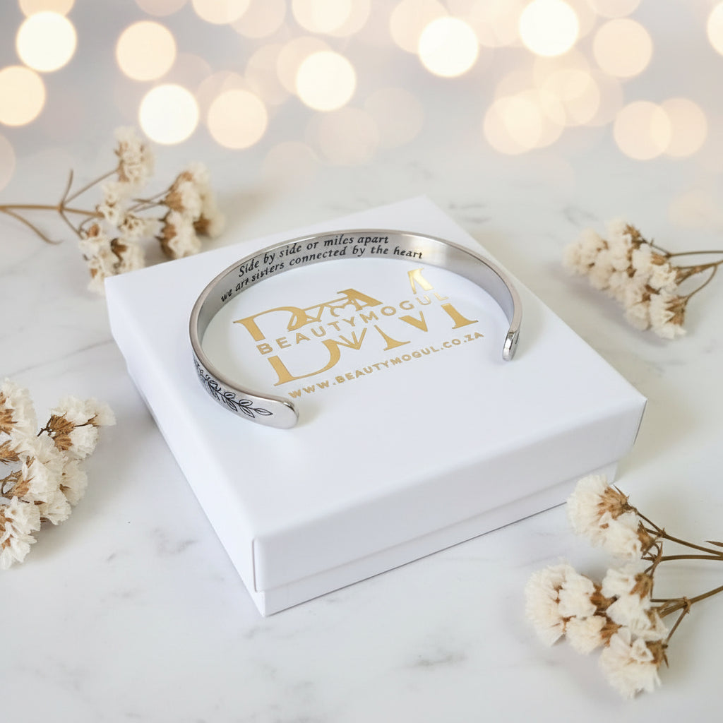 A silver-toned engraved cuff bracelet displayed on a white gift box, surrounded by dried white flowers on a light marble surface with soft bokeh lights in the background.