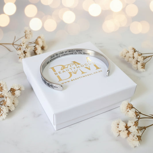 A silver-toned engraved cuff bracelet displayed on a white gift box, surrounded by dried white flowers on a light marble surface with soft bokeh lights in the background.