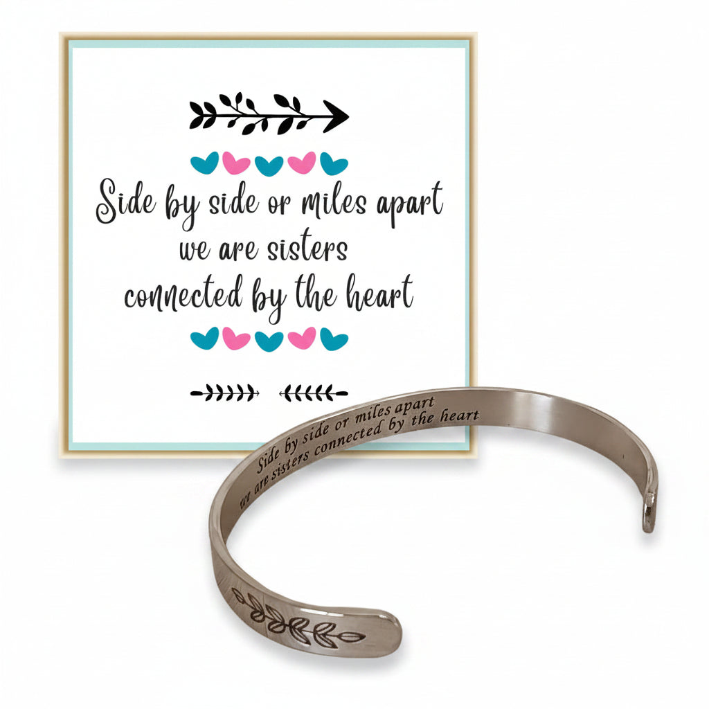 A silver-tone open cuff bracelet with engraved text and leaf motifs, shown in front of a matching printed sign on a white background. The sign reads, 'Side by side or miles apart we are sisters connected by the heart' with small blue, pink, and black heart accents.
