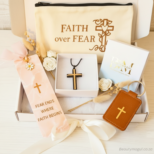 A curated gift set arranged in an open white box on a light wood surface. The set includes a cream canvas pouch printed with 'FAITH over FEAR,' a pink ribbon bookmark with a gold cross and text, a black-and-gold cross pendant in a small white box, a white branded box, a brown leather keychain with a gold cross, dried flowers, and decorative ribbons.