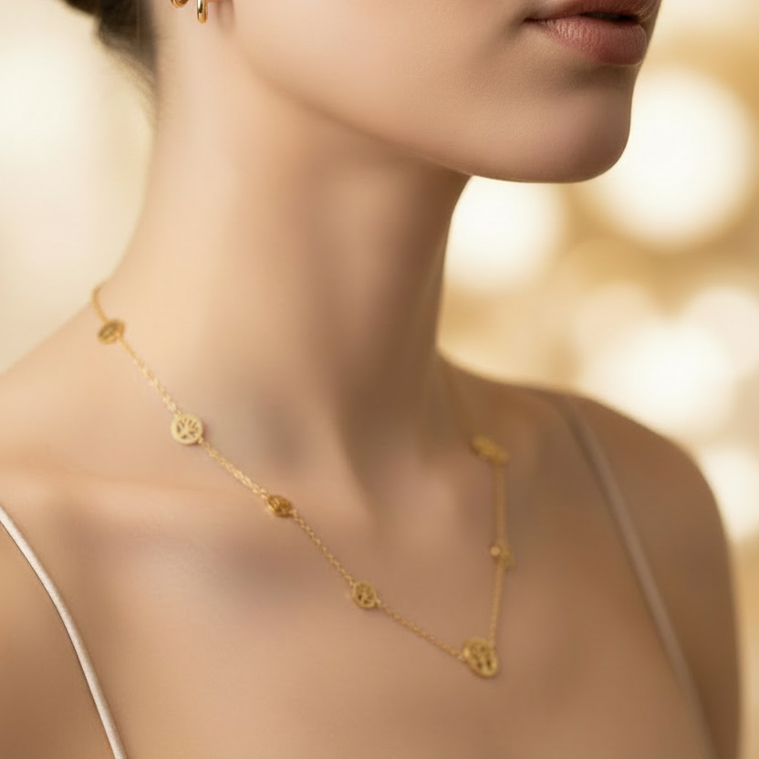 A close-up lifestyle shot of a model wearing a delicate gold necklace with small circular medallions and matching gold hoop earrings, styled with a satin cream top against a warm, softly blurred background.