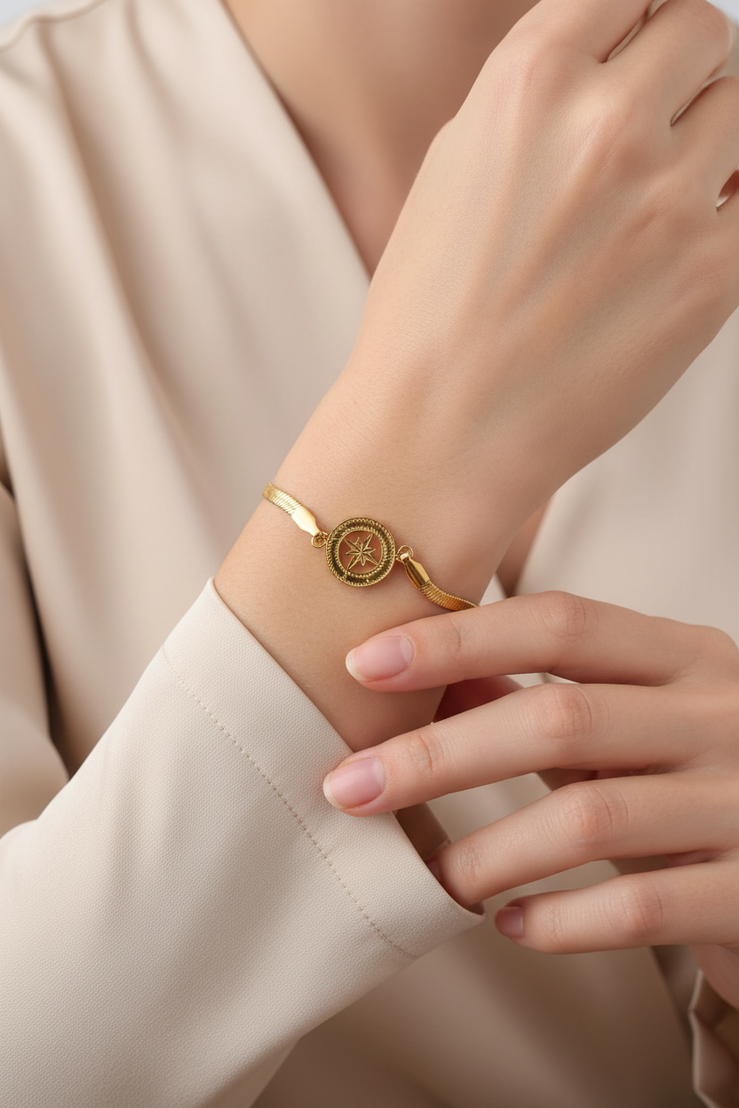 A close-up of a person's wrist wearing a delicate gold bracelet with a circular compass design, styled with a soft beige blouse.
