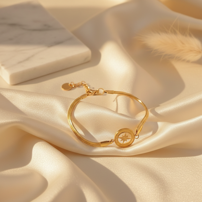 A gold-toned snake chain bracelet with a circular compass design, styled on draped champagne satin fabric with a marble block and dried grass prop.