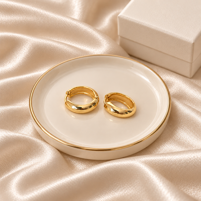 Two polished gold hoop earrings displayed on a small round white tray with a gold rim, set on draped champagne satin fabric with a white jewelry box in the background.