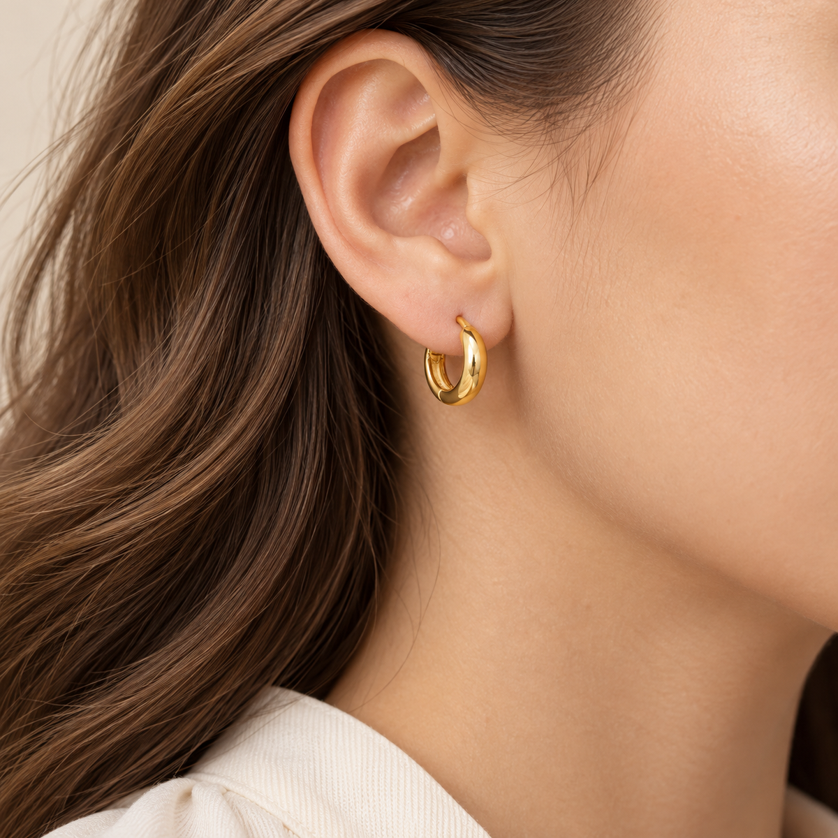 A close-up of a woman's ear wearing a small polished gold huggie hoop earring.