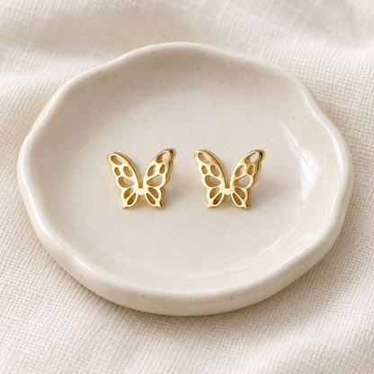 A pair of gold butterfly stud earrings displayed on a small scalloped cream ceramic dish over a textured beige fabric background.