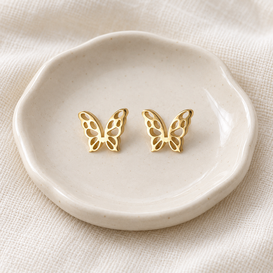 A pair of gold butterfly stud earrings displayed on a small scalloped cream ceramic dish over a textured beige fabric background.