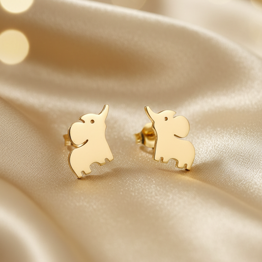 A pair of small gold elephant stud earrings resting on draped champagne satin fabric with soft golden bokeh lights in the background.